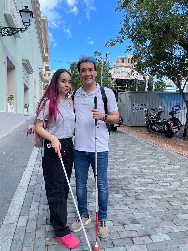 En la foto hay un hombre y una mujer parados en una calle empedrada. Ambos tienen bastones blancos para ciegos.  La mujer tiene el cabello largo y teñido de rosa, lleva una camiseta gris, pantalones negros y zapatos rosas. El hombre tiene el cabello corto y grisáceo, lleva una camiseta blanca, jeans azules y zapatos marrones. Lleva una mochila negra y sonríen a la cámara. Detrás de ellos hay edificios de colores claros y un letrero que dice "SAN SEBASTIAN" en el fondo. Hay motocicletas e